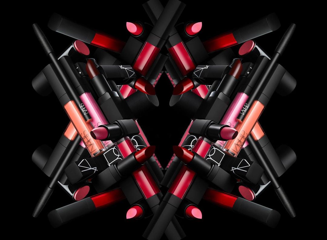 Close-up product shot of an arranged assortment of NARS brand lipsticks, lip pencils, and lip gloss tubes against a black background.