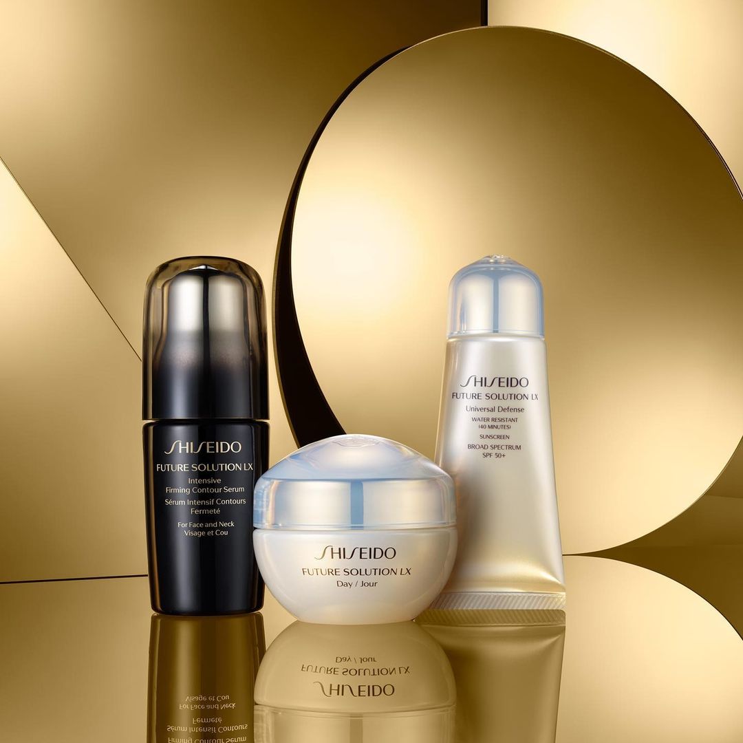 Three Shiseido Future Solution LX skincare product containers sit on a reflective surface with a golden patterned background. Beauty shot.