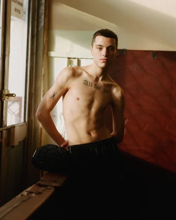 Shirtless young man standing indoors near a window with gothic lettering tattoo across his chest, photographed by Brett Lloyd.