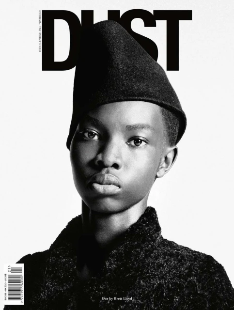 Black and white portrait of African female fashion model wearing a dark colored hat and coat against a plain white studio backdrop.