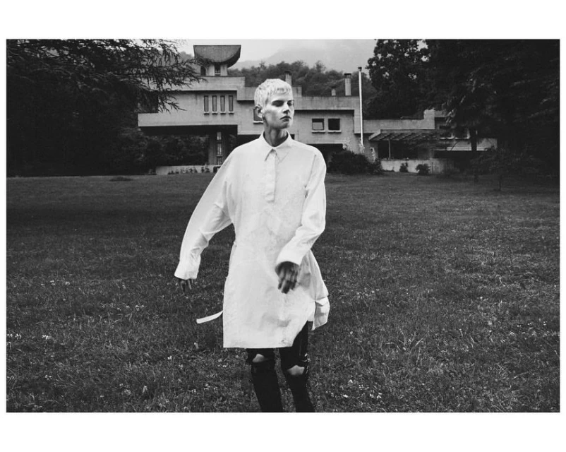 Monochrome shot of a person with a buzzcut wearing a long white shirt and black ripped jeans walking in a grassy yard with a building.