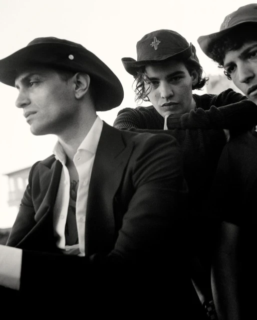 Three young men in vintage hats and clothing pose in an undated black and white photograph with a bright, out of focus background.