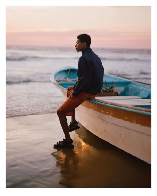 A young man sits on a blue and white boat at the beach at dusk, wearing a dark jacket, orange shorts, and sandals, with waves in background.