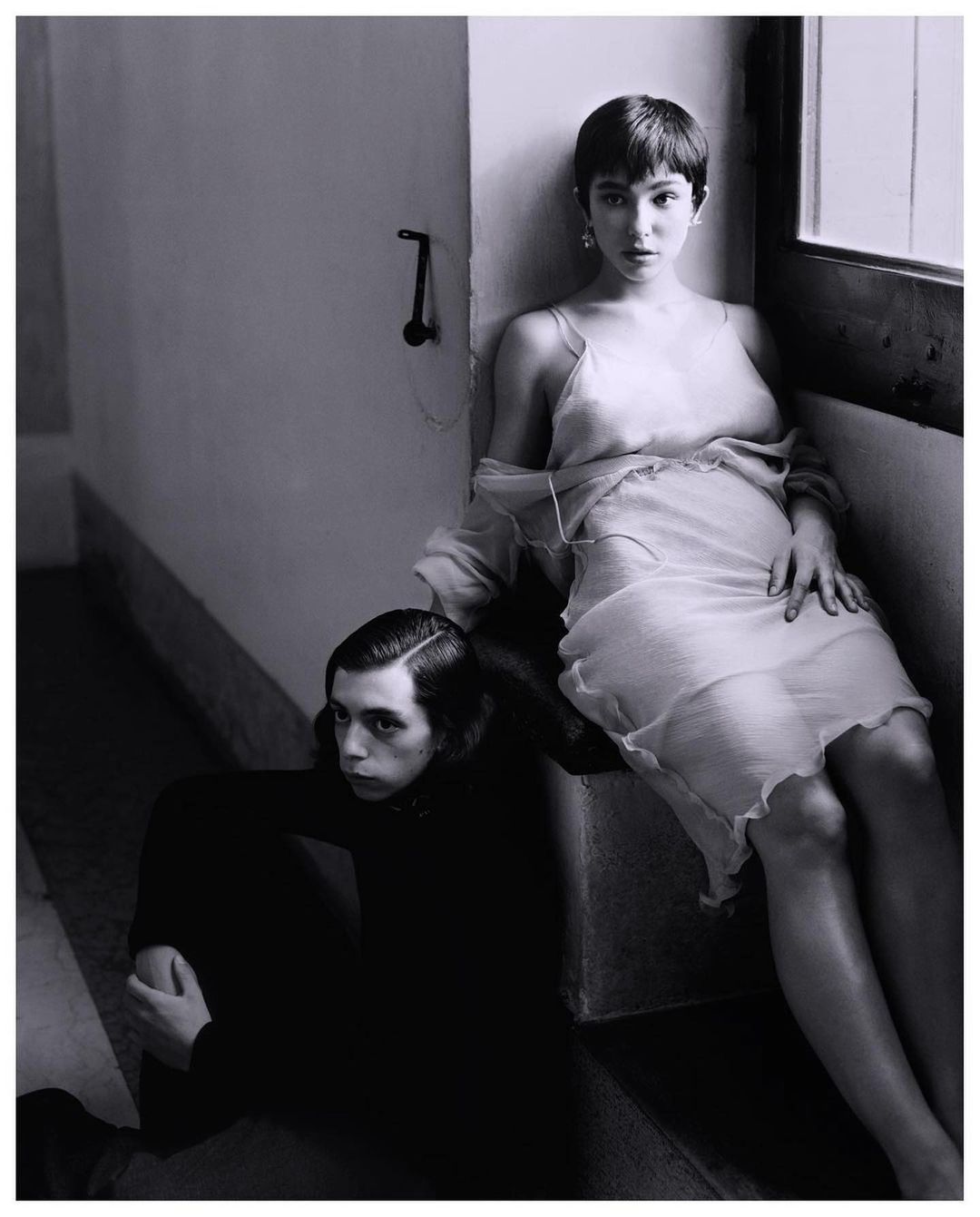 A young woman in a semi-sheer dress seated on a step next to a young man sitting on the floor in a neutral tone, indoor fashion shot.