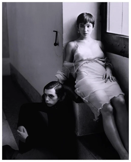 A young woman in a semi-sheer dress seated on a step next to a young man sitting on the floor in a neutral tone, indoor fashion shot.