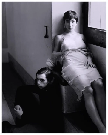 A young woman in a semi-sheer dress seated on a step next to a young man sitting on the floor in a neutral tone, indoor fashion shot.