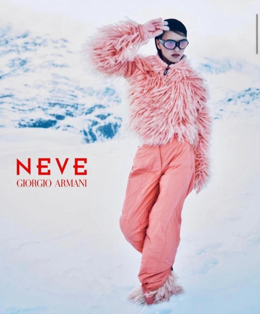 A woman poses in the snow wearing a pink fur jacket, snow goggles, and matching pants, against a snowy mountain backdrop.