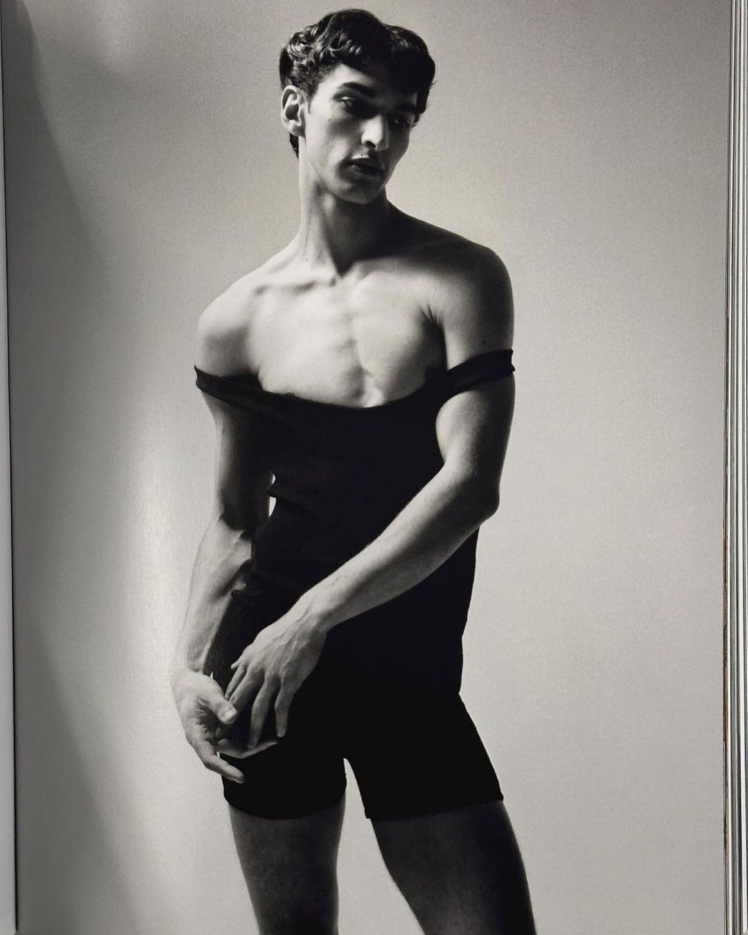 A slender man in a black bodysuit strikes a pose in a studio portrait with soft lighting against a light-grey background.