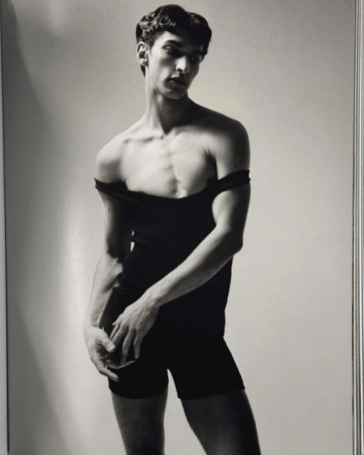A slender man in a black bodysuit strikes a pose in a studio portrait with soft lighting against a light-grey background.