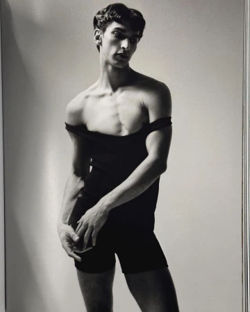 A slender man in a black bodysuit strikes a pose in a studio portrait with soft lighting against a light-grey background.