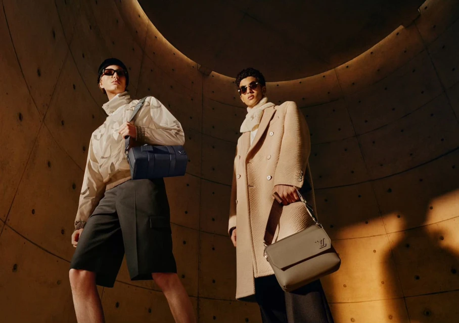 Two adult male models pose in a tan concrete tunnel wearing sunglasses, modern outerwear, and carrying handbags in a fashion editorial.