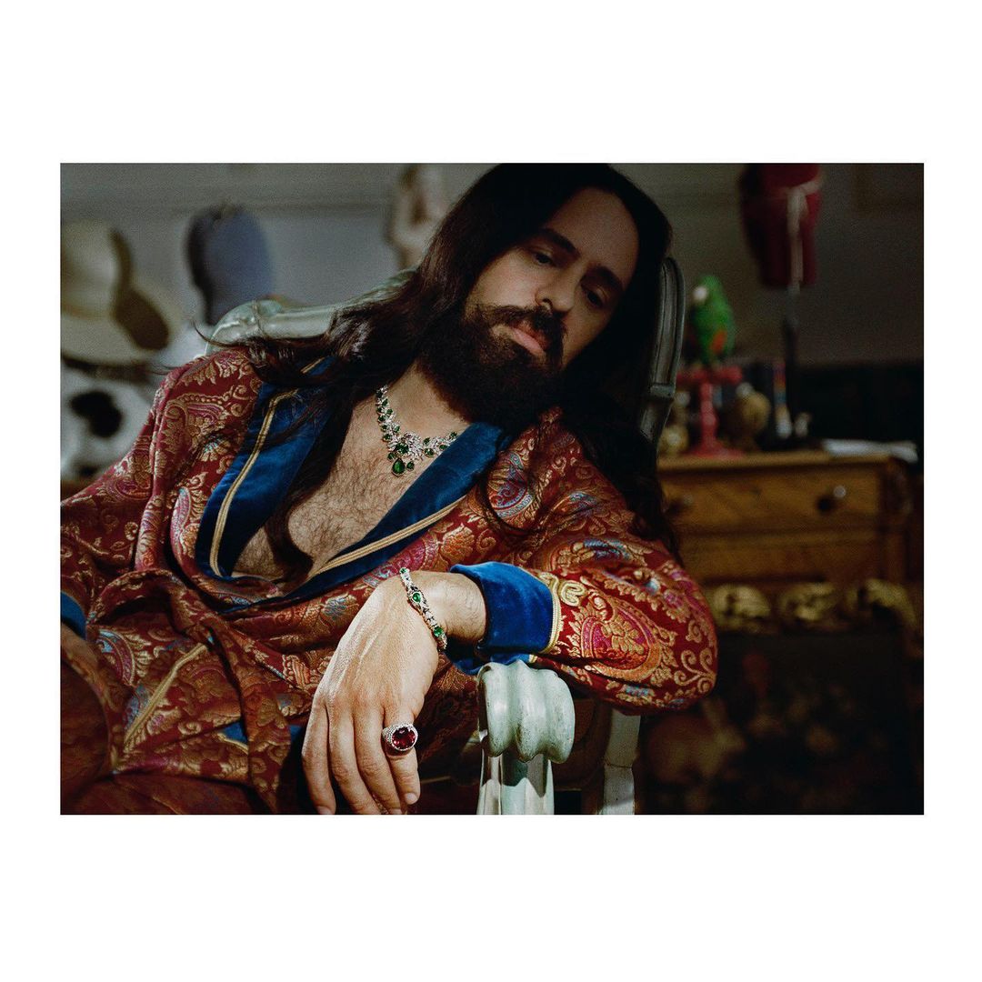 Man in paisley robe with beard and long hair sits in a chair with jewelry, indoors, with hats, drawers, and a parrot statue in the background.