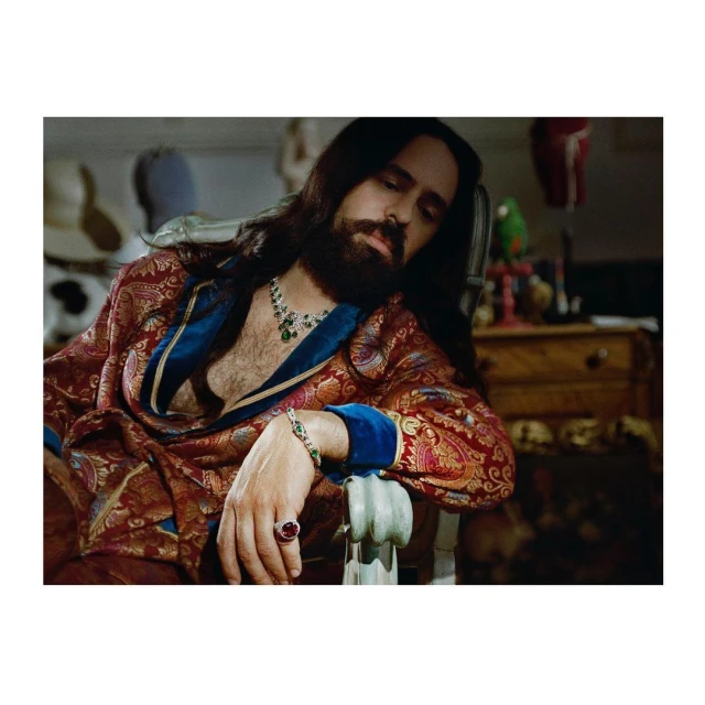 Man in paisley robe with beard and long hair sits in a chair with jewelry, indoors, with hats, drawers, and a parrot statue in the background.