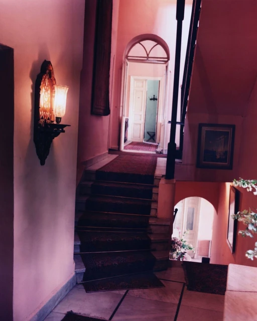 Interior view of a pink-toned staircase with a decorative wall lamp, leading to an arched doorway and a glimpse of an adjacent room.
