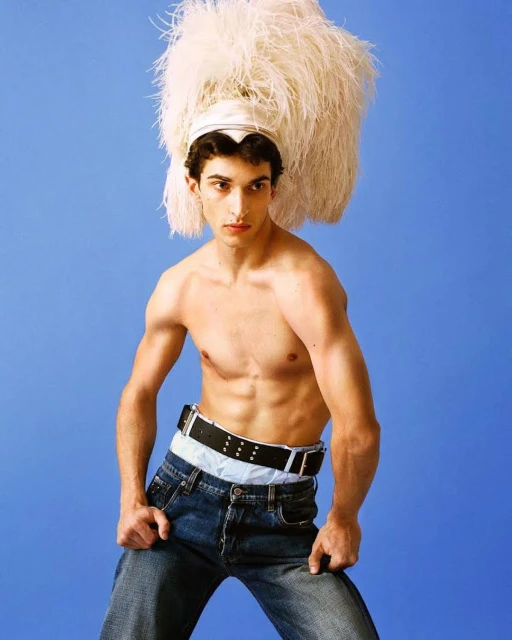 Shirtless man poses against a blue backdrop wearing jeans, a light blue top and black belts. He wears an elaborate feathered hat.