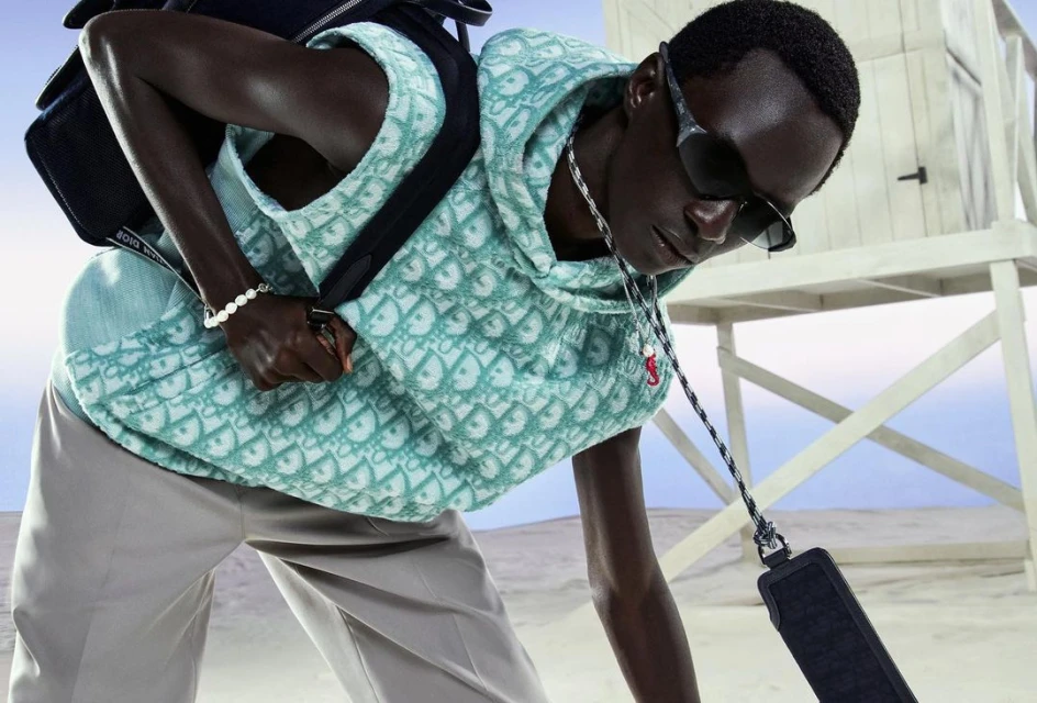 Masculine fashion shoot: A man wears a patterned teal hoodie vest, sunglasses, and light pants on the sand with a white lifeguard tower.