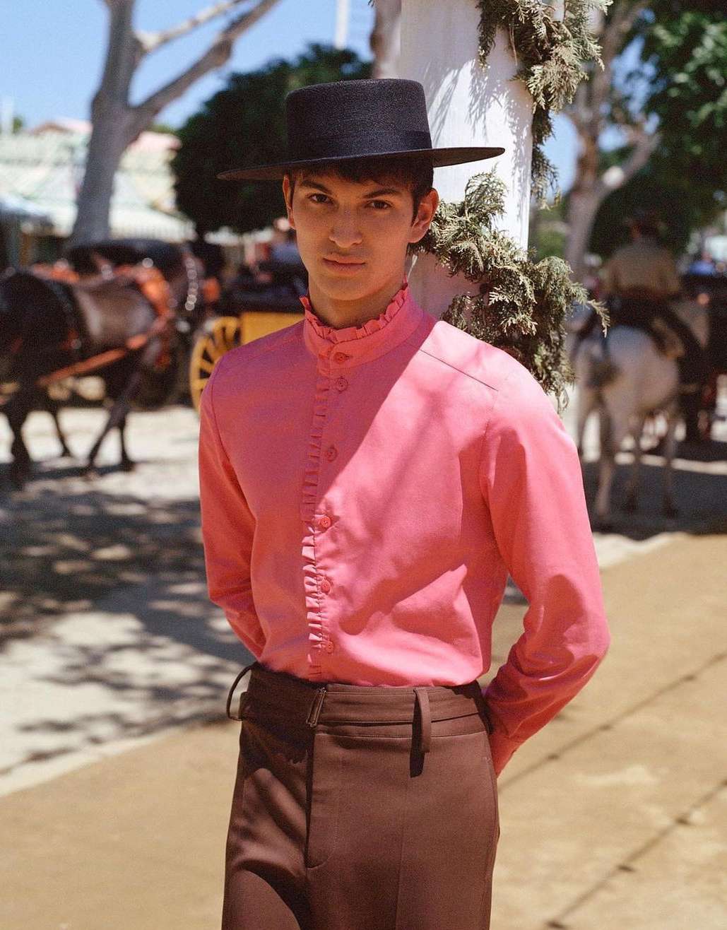 Young adult male wearing a traditional hat and pink ruffled shirt poses outdoors near horses and carriages in a sunny environment.