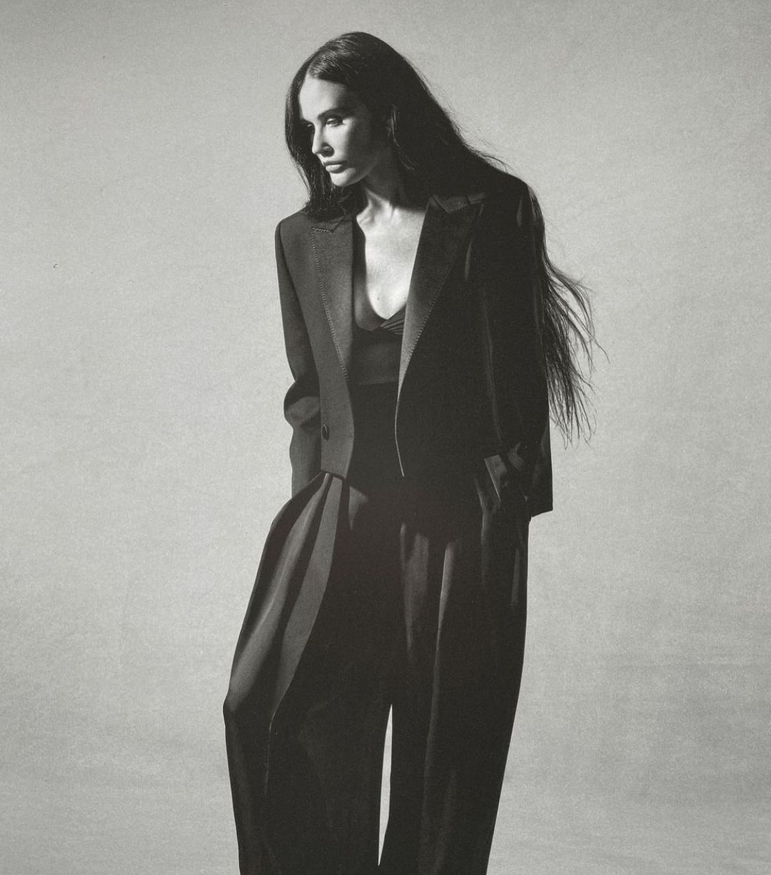 Elegant adult woman with long dark hair poses in a stylish black blazer and palazzo pants against a neutral grey backdrop in a studio shot.