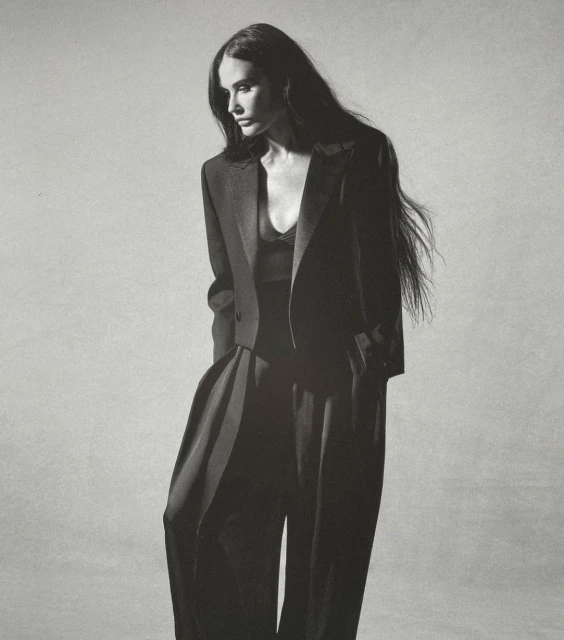Elegant adult woman with long dark hair poses in a stylish black blazer and palazzo pants against a neutral grey backdrop in a studio shot.