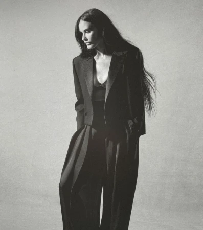 Elegant adult woman with long dark hair poses in a stylish black blazer and palazzo pants against a neutral grey backdrop in a studio shot.