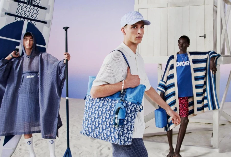 Three fashion models in beachwear accessories and a tote bag promote a high-end brand on a sandy beach with a white lifeguard station.