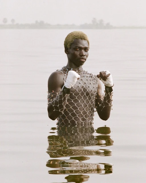 Adult male in water with boxing wraps and net shirt; outdoors, eye-level shot with neutral color palette and view of distant trees.