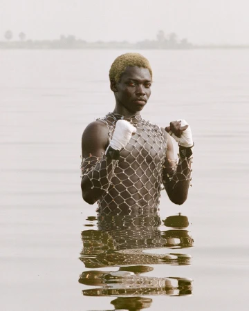 Adult male in water with boxing wraps and net shirt; outdoors, eye-level shot with neutral color palette and view of distant trees.
