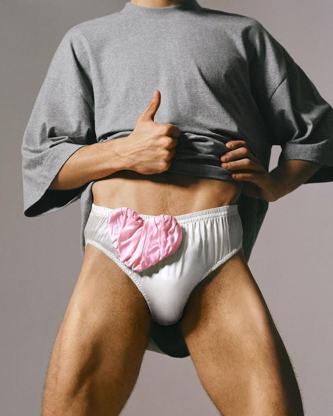 Torso shot of an adult male wearing white briefs with a pink heart, an oversized grey tee-shirt, and giving a thumbs-up gesture.
