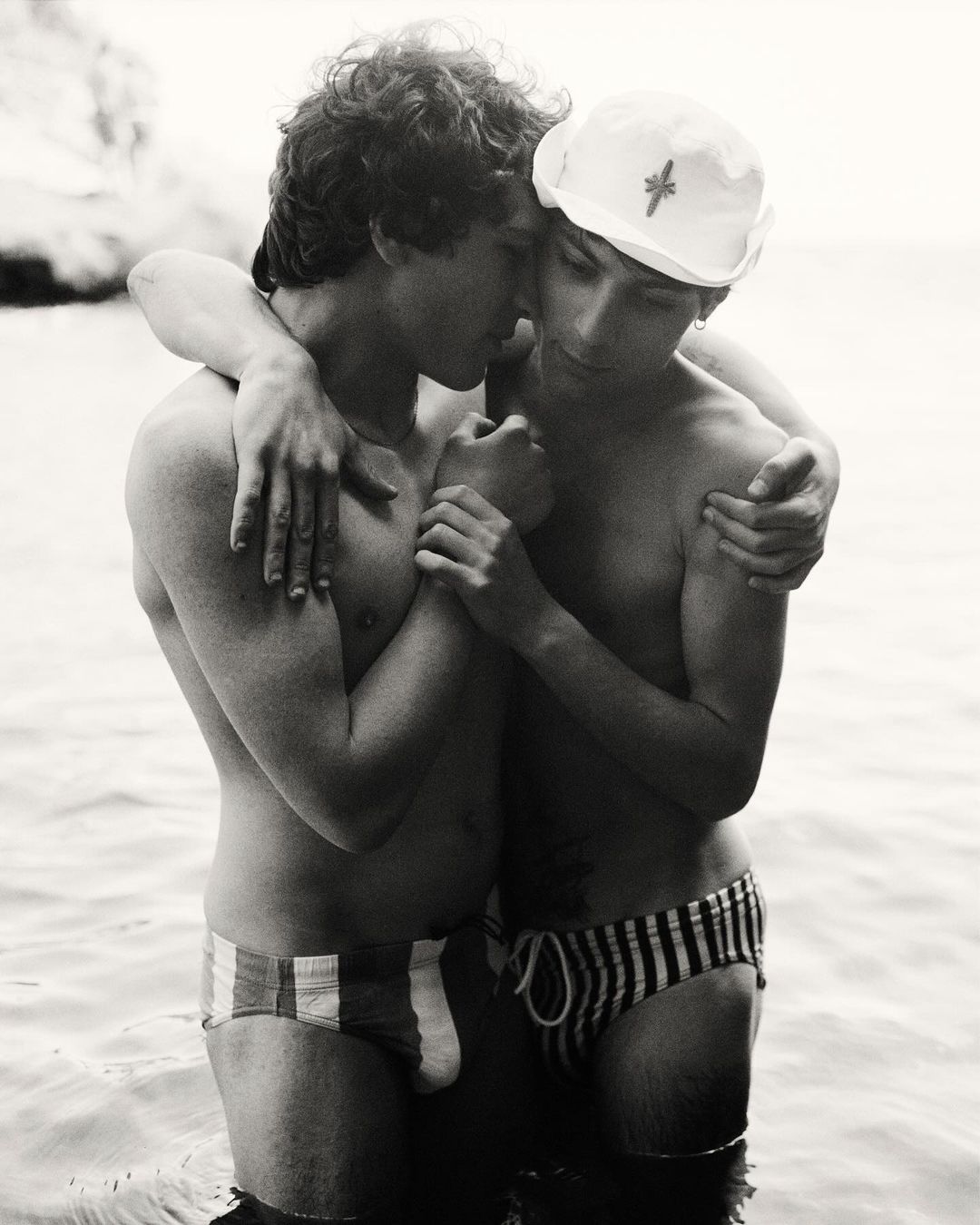 Two young adult males embracing in shallow water, wearing swim briefs, against a blurred coastal background in a black and white photo.