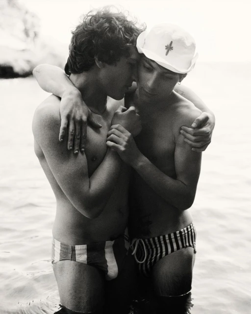 Two young adult males embracing in shallow water, wearing swim briefs, against a blurred coastal background in a black and white photo.