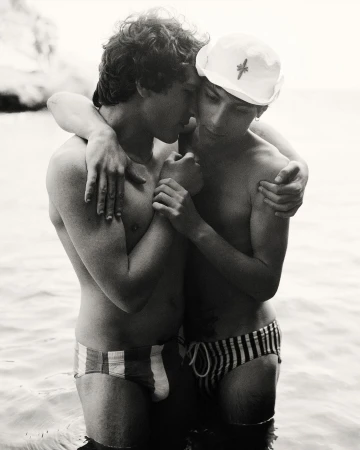 Two young adult males embracing in shallow water, wearing swim briefs, against a blurred coastal background in a black and white photo.