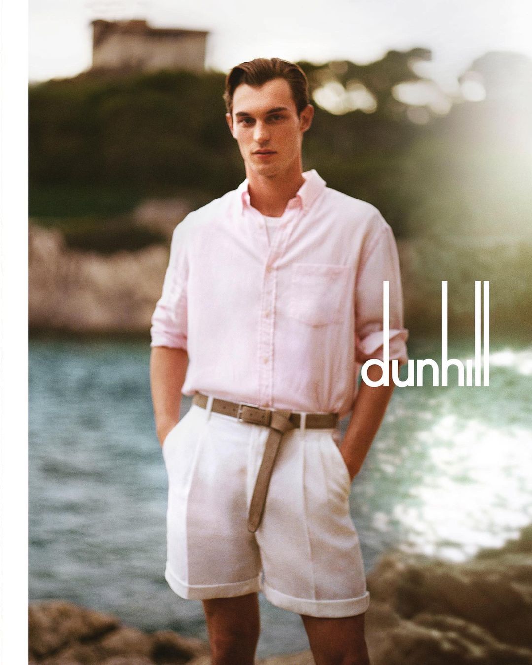Fashion shot of a single male model in a pink shirt and white shorts, standing near the ocean with an island in the background.