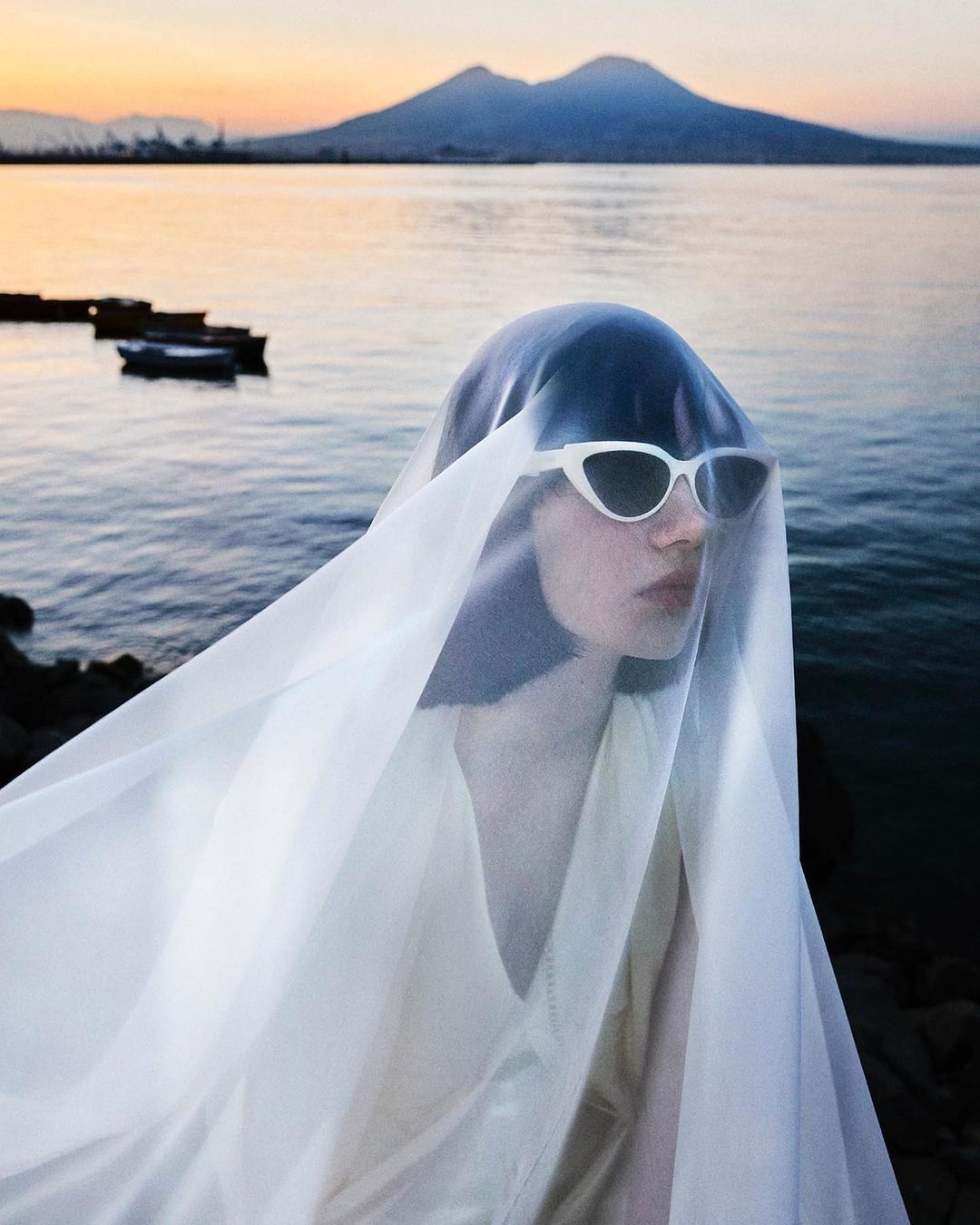 Woman wearing white cat eye sunglasses and a sheer white veil over her head by the ocean at sunset.