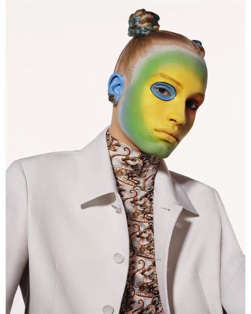 Portrait of a model with green and yellow face paint, blue painted ears, and stylized hair, wearing a white jacket and patterned shirt.