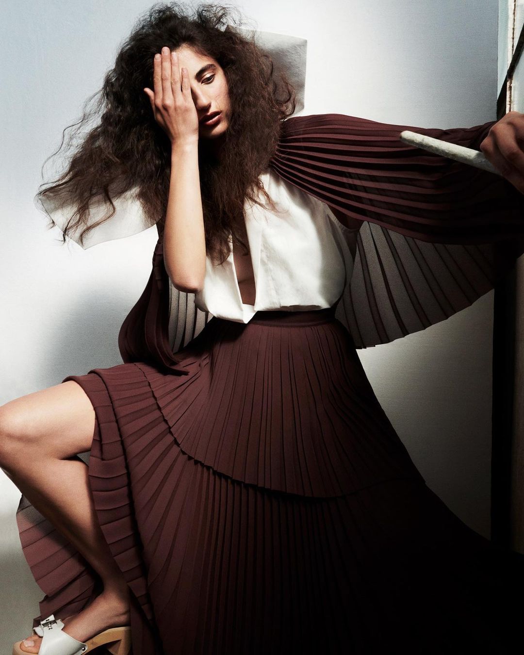 Fashion shot of woman with curly hair in a pleated skirt and white top, hand covering eye, sitting in front of a neutral wall.