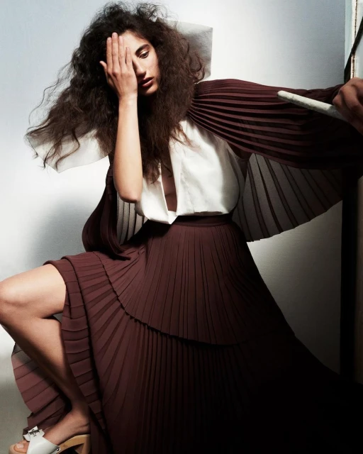 Fashion shot of woman with curly hair in a pleated skirt and white top, hand covering eye, sitting in front of a neutral wall.
