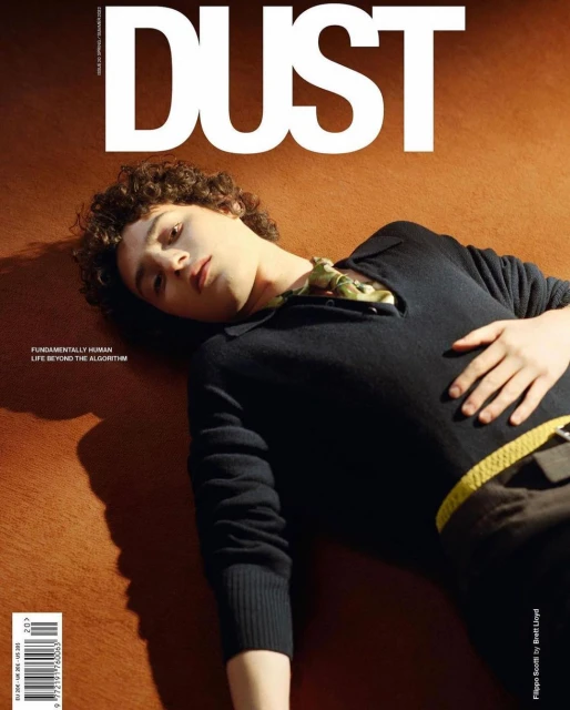 Magazine cover featuring a male model with curly hair, laying on a bronze backdrop, wearing a black v-neck sweater and brown pants.