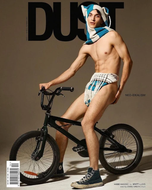 Shirtless male fashion model on a black BMX bike wearing a striped white and blue hat and fringed garment for DUST Magazine.