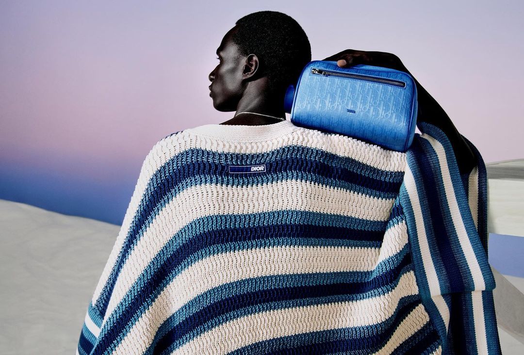 Man wearing a blue and white striped sweater and holding a Dior toiletry bag over his shoulder, posing in front of a gradient background.