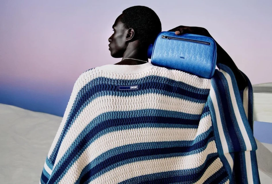 Man wearing a blue and white striped sweater and holding a Dior toiletry bag over his shoulder, posing in front of a gradient background.