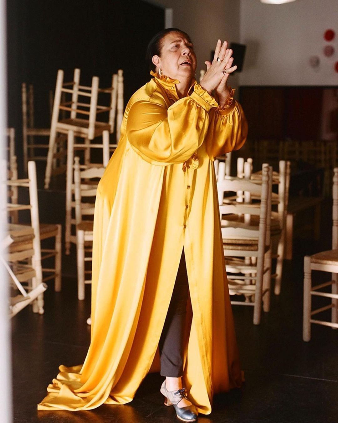 An adult woman wearing a long yellow robe stands on a black floor in front of stacked chairs clapping her hands together.
