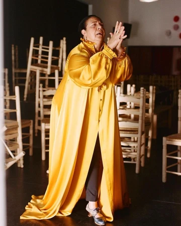 An adult woman wearing a long yellow robe stands on a black floor in front of stacked chairs clapping her hands together.