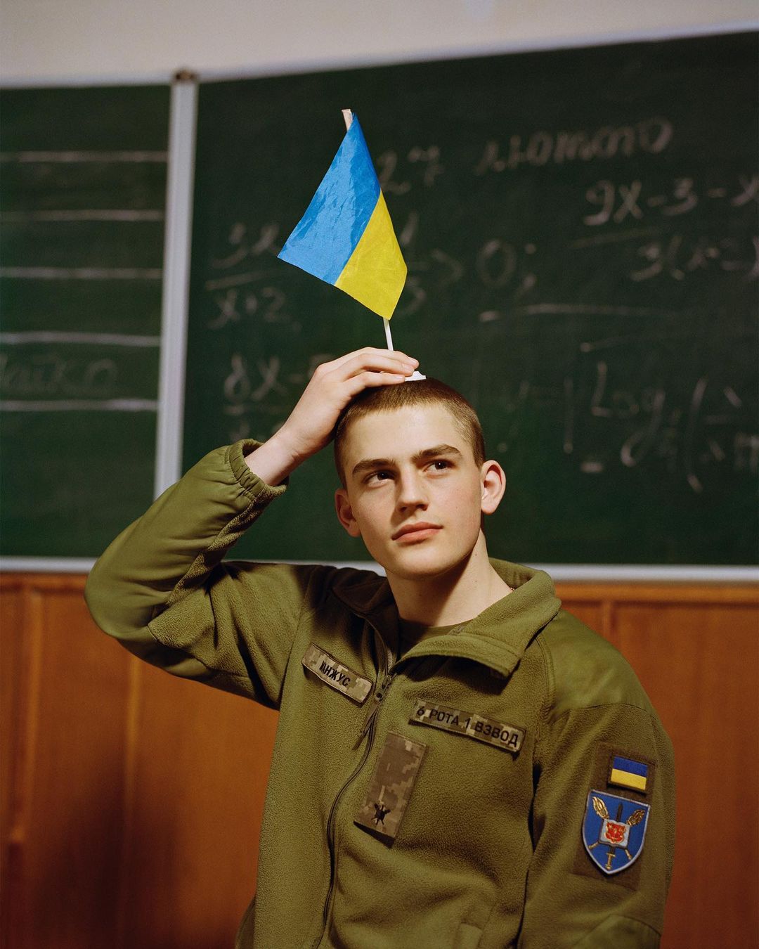 Young man in military jacket poses with a Ukrainian flag on his head in a classroom, looking thoughtfully to the side.