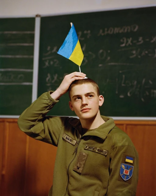 Young man in military jacket poses with a Ukrainian flag on his head in a classroom, looking thoughtfully to the side.