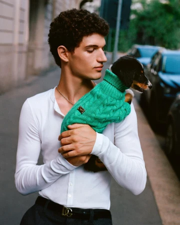 Young man with curly dark hair wearing a white long-sleeve henley holding a small puppy in a bright green cable-knit sweater.