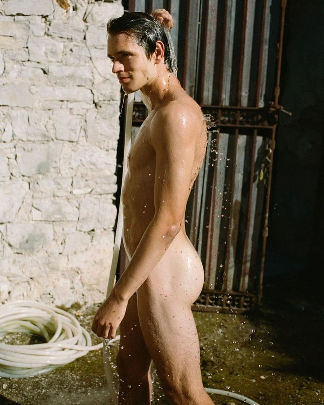 Nude male showering outdoors with a hose against a weathered stone wall and metal door, bathed in warm light.