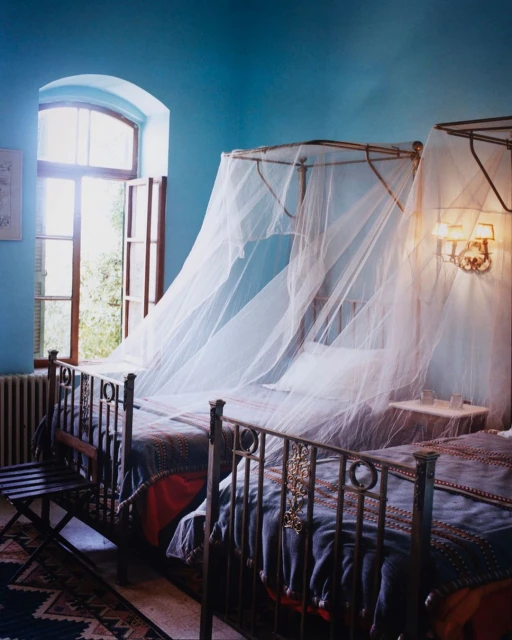Bedroom interior featuring two wrought iron canopy beds draped with mosquito netting near an open window with blue walls.