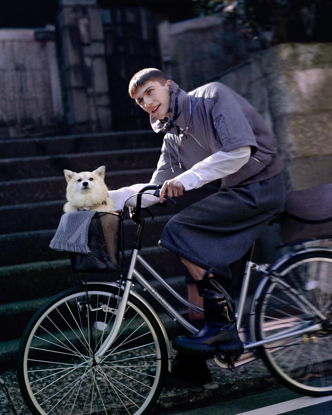 Man riding a bicycle with a fluffy white dog in the front basket on city street, wearing a jacket, shorts and boots.