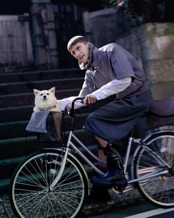 Man riding a bicycle with a fluffy white dog in the front basket on city street, wearing a jacket, shorts and boots.