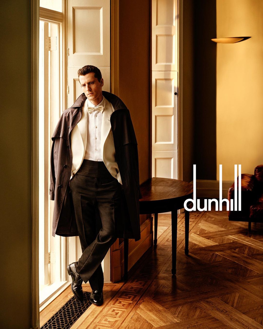 Elegant man in a trench coat poses next to a sunlit window, showcasing a round wooden table and the 'dunhill' logo on a parquet floor.